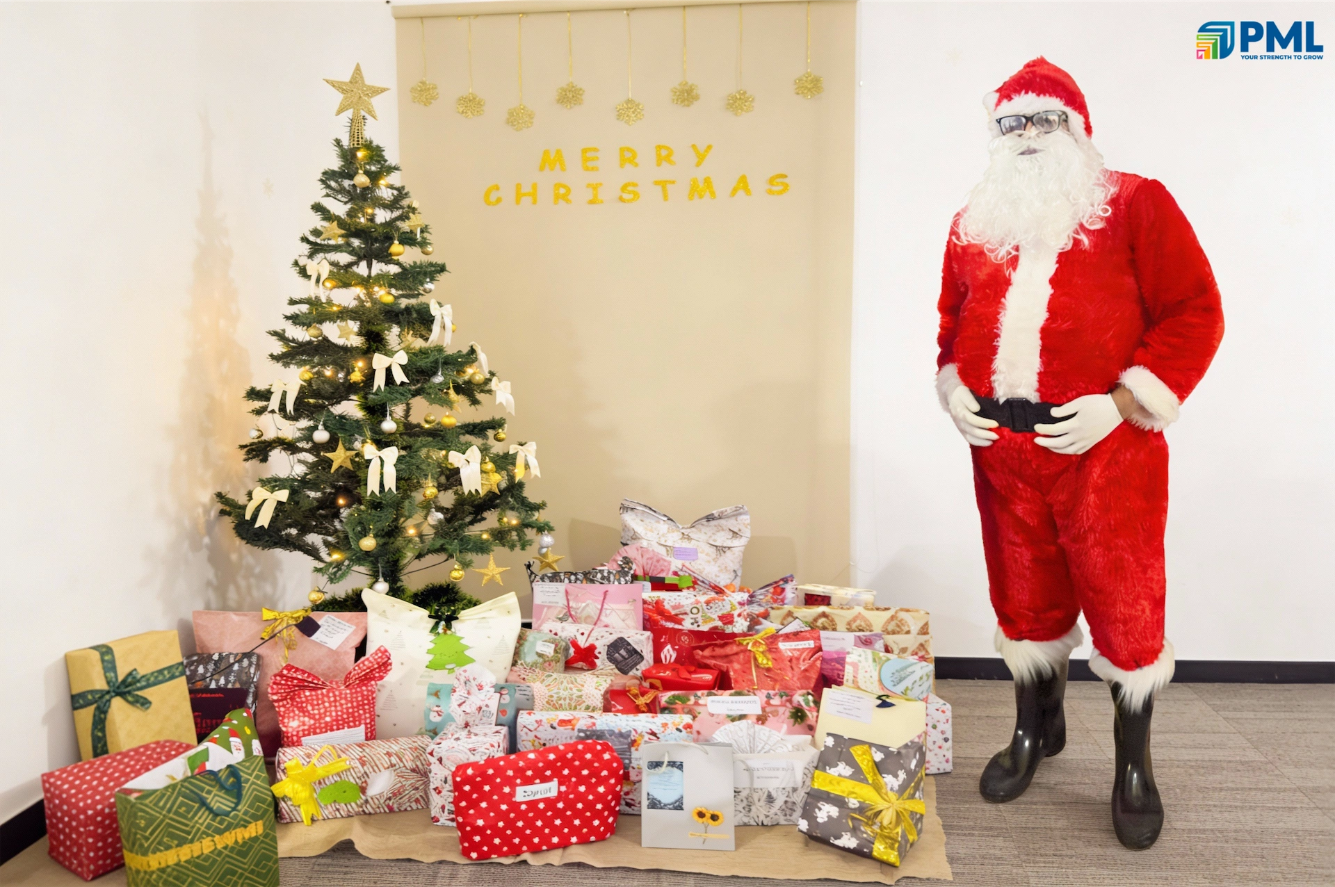 Person dressed as Santa Claus standing beside a Christmas tree and gift boxes in an office setting
